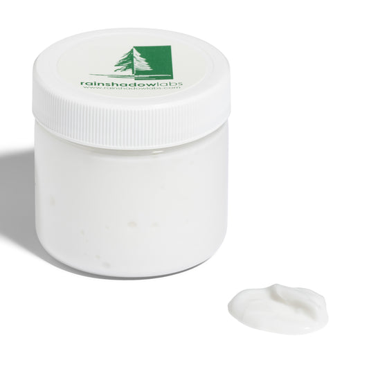 A white jar of Shave Cream with a white ribbed lid, branded with the Rainshadow Labs logo on the lid. A small amount of the lotion is applied to a cotton pad beside the jar.